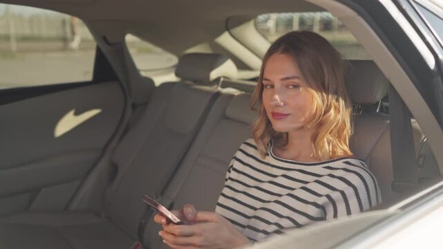 Young beautiful woman sitting in back seat of car, holding smartphone with both hands and smiling. Looking at screen with slight smile and chatting on mobile phone. Female passenger with gadget.