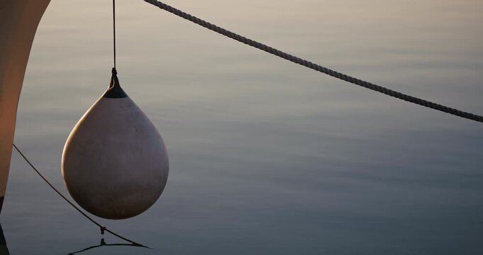 White boat fender hanging from a docked boat above calm water with mooring rope in low sunlight.