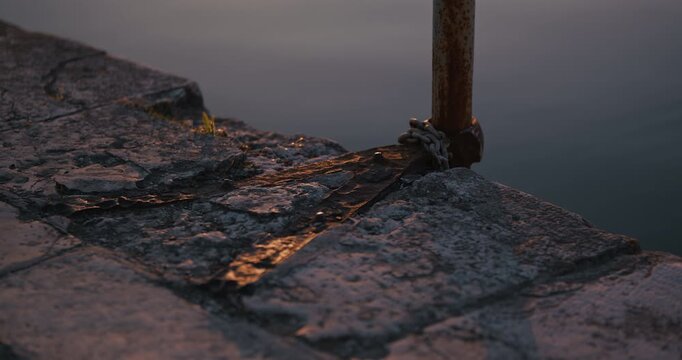 Rusty metal post with chain mounted on stone waterfront edge above calm water in low sunlight.