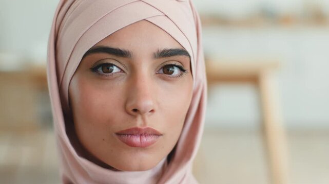 Close up pensive Muslim woman in pink hijab looking at camera serious indoor portrait Arabian female calm face Islamic girl thoughtful gaze ethnic lady model headscarf headwear fashion beauty indoors
