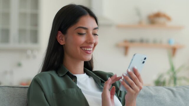 Happy smiling Indian Muslim woman using smartphone at home sofa shopping online ordering food delivery scrolling social media app relaxed Arabian female girl browsing mobile phone internet indoors