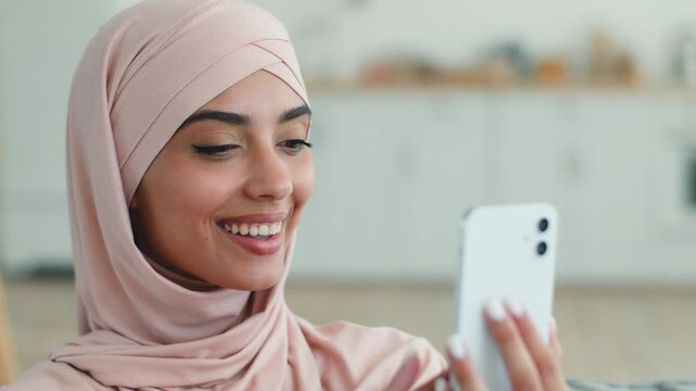 Excited amazed happy Indian Arabian Muslim woman in hijab smiling at mobile phone indoor female Islamic girl say wow celebrate victory good news online success achievement smartphone at home apartment