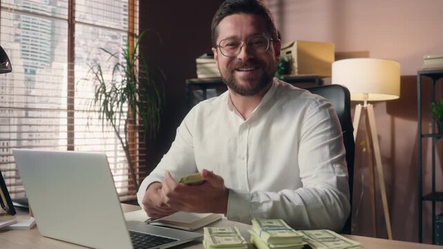 Glad smiling Caucasian business man holding stack cash indoor office CEO leader smelling dollars banknotes earnings laptop businessman entrepreneur salesman male financial growth money wealth triumph