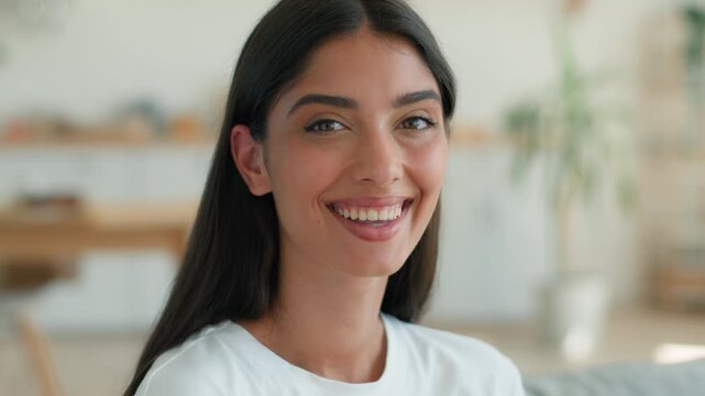 Happy smiling Indian Arabian Muslim woman looking at camera on sofa at home indoors positive carefree female girl wife toothy healthy smile relaxing on couch apartment living tenant mortgage portrait