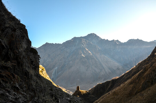 A lone, ancient watchtower stands silhouetted in deep mountain valley. Golden sunlight catches the rugged peaks and slopes of the Caucasus range under clear, pale sky in this serene landscape. Georgia