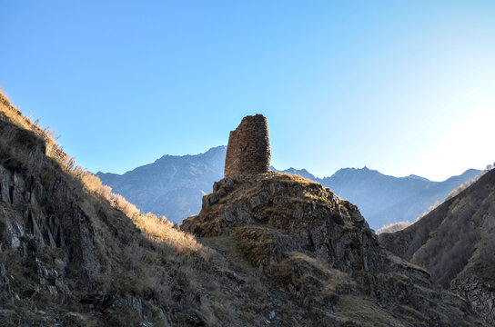 A lone, ancient watchtower stands silhouetted in deep mountain valley. Golden sunlight catches the rugged peaks and slopes of the Caucasus range under clear, pale sky in this serene landscape. Georgia