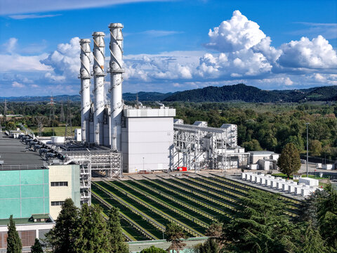 Aerial view of the A2A Chivasso Power Plant featuring three tall industrial chimneys and modern facilities surrounded by green hills and trees in Chivasso, Piemonte, Italy.