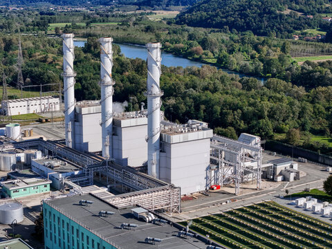 Aerial view of the A2A Chivasso Power Plant featuring three tall white chimneys and industrial structures near the river and green hills in Chivasso, Piemonte, Italy.