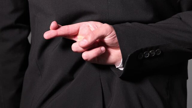 Man in black suit crossing fingers behind his back on grey background, closeup. Camera moving forward