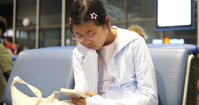 Cinematic close-up of a young Asian girl with glasses using a smartphone while waiting at an airport terminal, travel lifestyle and digital technology concept