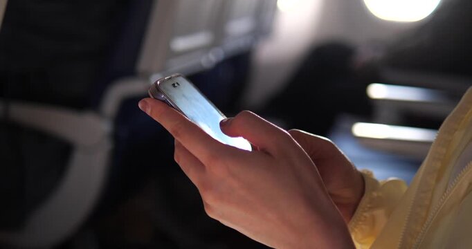 Cinematic close-up of a passenger using a smartphone on an airplane, bright screen light reflecting on hands in a dark cabin, travel technology concept