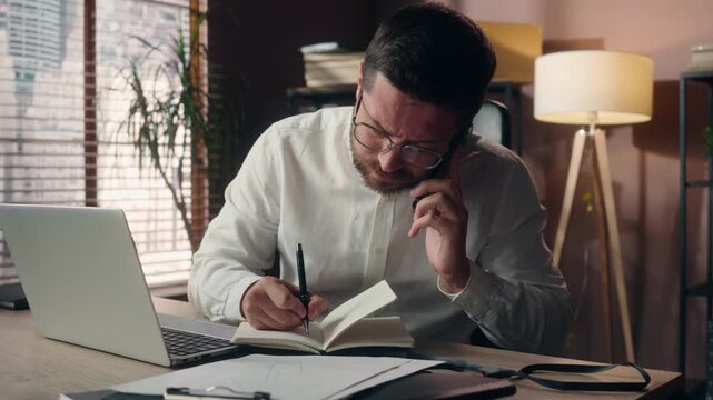 Overworked Caucasian business man multitasking in office on mobile phone writing notes using laptop stressed businessman manager executive entrepreneur deadline cellphone overload male worker indoors