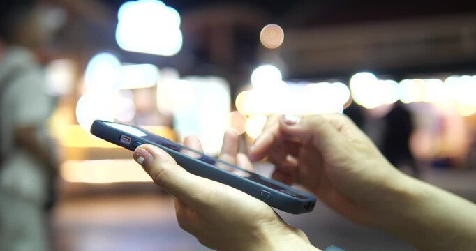 Cinematic close-up of hands using a smartphone on a city street at night, beautiful colorful bokeh background and urban lifestyle concept