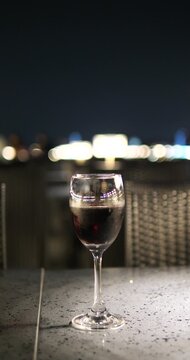 Cinematic close-up of a red wine glass on a table at night, luxury cruise or rooftop bar atmosphere with elegant lamp and city bokeh background