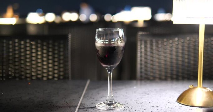 Cinematic close-up of a red wine glass on a table at night, luxury cruise or rooftop bar atmosphere with elegant lamp and city bokeh background