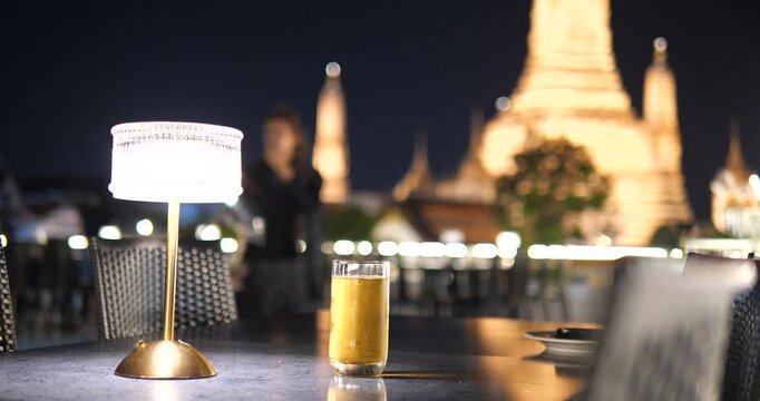 Cinematic close-up of a cold beer glass on a table at night, luxury riverside dining or cruise atmosphere with blurred Wat Arun Temple background in Bangkok, Thailand