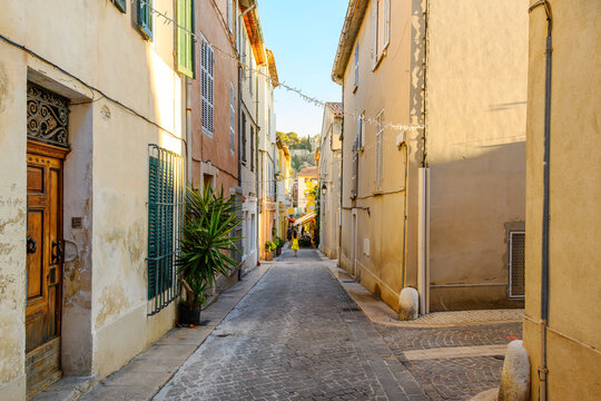 A young woman walks along a narrow residential cobblestone street of shops and sidewalk cafes towards the old town section of the seaside town of Cassis, France, French Riviera.