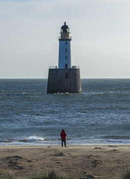 Aerial view of Rattray Head Lighthouse standing in the North Sea with a person in a red jacket on the sandy beach in the foreground in Peterhead, Scotland, United Kingdom.