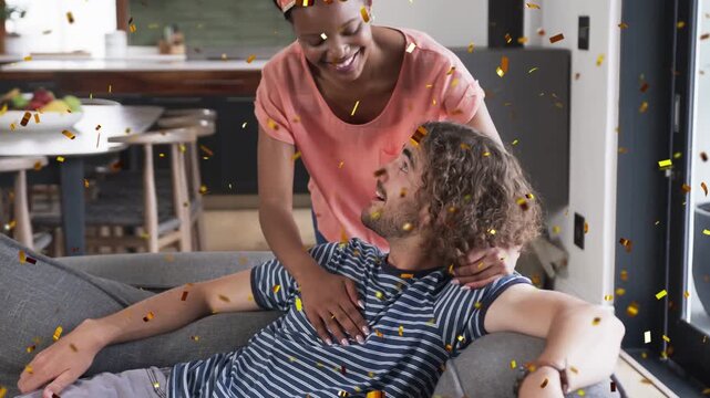 Man reclining on gray sofa, woman walking up causing metal confetti falling, couple embracing home