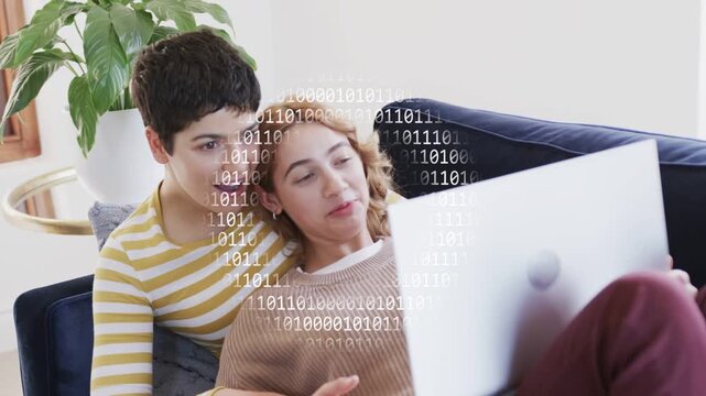 Two women cuddling on couch clicking laptop together and viewing binary-code overlay for technology