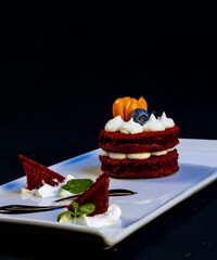 Elegant plated red velvet cake with cream cheese frosting, blueberries, physalis and mint leaves on a white plate. Black background. Fine dining dessert concept. © Aziza