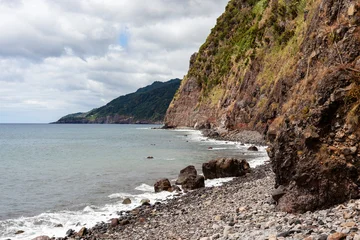 Fototapeten Küste Rocky coastline near Povoacao on Sao Miguel Island. The rugged cliffs meet the Atlantic Ocean's waves. Green vegetation covers the steep hillsides under a cloudy sky.  © Eugene Ga