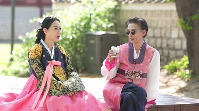Two women in traditional Korean attire sit outside. One wears bright pink with gold embroidery. The other wears pink with a patterned belt. They smile, engaged in light conversation