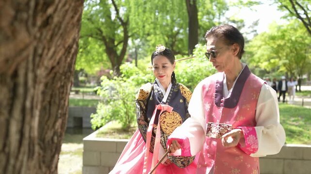 Two people in traditional Korean dress stroll under trees. They smile and share a quiet moment together. Bright green foliage frames their gentle walk. Ideal for cultural, wedding, or travel projects
