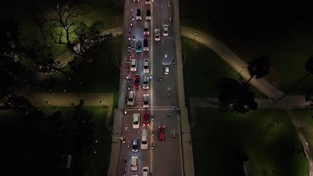 Night Bridge In Hue City, Asian Commuters Stuck In Slow Traffic On Narrow Lanes, Aerial Top View Of Long Queue Of Cars And Motorcycles Alongside Dark River Bank And Lit Park.