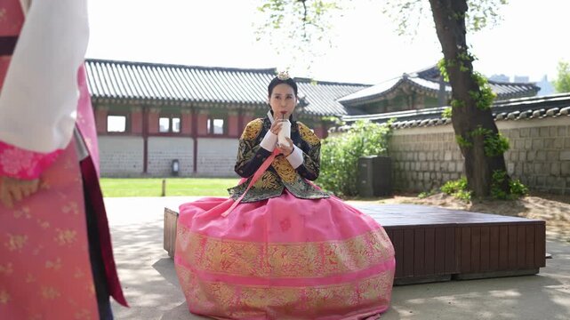 Woman in traditional Korean attire sits gracefully. She wears a vibrant pink skirt with gold patterns. Man stands nearby, dressed in matching traditional wear. He leans forward slightly