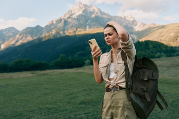 Obraz premium A woman outdoors checks her phone while wearing a hiking backpack, framed by rugged mountains, capturing a spontaneous travel lifestyle moment on a bright day with open fields.