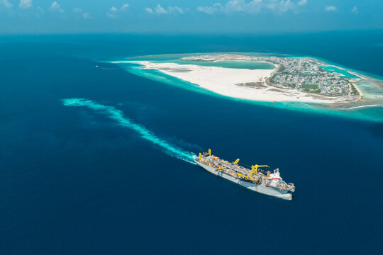 Aerial view of a large industrial dredger ship sailing through deep blue ocean waters towards a tropical island with white beaches in Vashafaru, Haa Alif Atoll, Maldives.