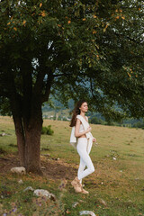 Obraz premium Woman lifestyle outdoors, standing near a tree in a sunlit meadow, wearing white outfit, relaxed pose, gentle breeze, natural colors, peaceful countryside.