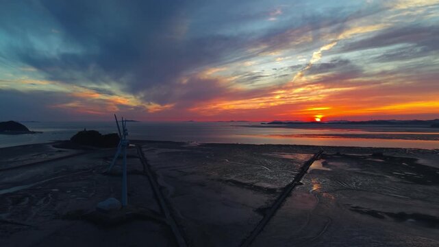 Camera pans left showing vibrant orange sunset over Tando Port mudflats with silhouettes of wind turbines and Nue Island at low tide