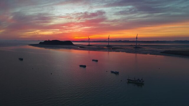 Low drone flypass over moored silhouetted trawler boats on glassy calm water approaching Nue Island Sea Road and three wind turbines at sunset