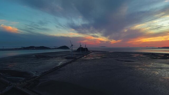 Cinematic wide shot of three large wind turbines on tidal mudflats during vibrant sunset along Tando Port Sea Road leading to Nue Island in Ansan South Korea
