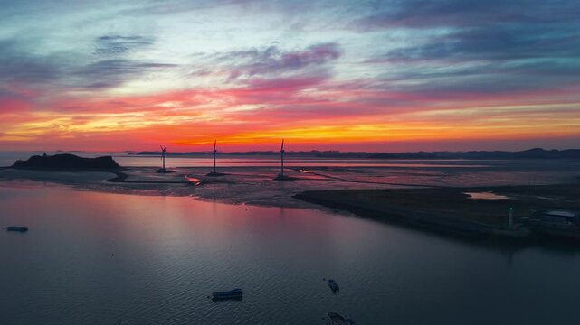 Cinematic aerial view of vibrant sunset at Tando Port with wind turbines silhouetted against orange sky over Nue Island sea road and anchored fishing boats in South Korea