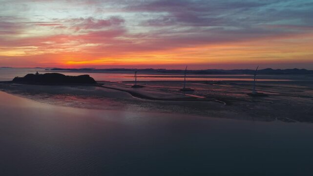 Aerial drone rises forward over exposed tidal mudflats of Nue Island Sea Road toward silhouetted wind turbines against vivid layered sunset sky