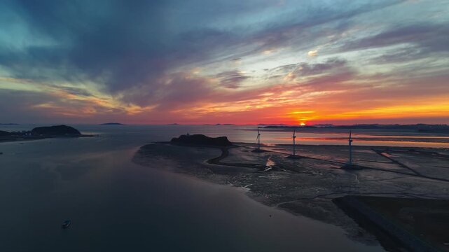 Aerial drone view of three silhouetted wind turbines on exposed tidal mudflats of Tando Sea Road with dramatic sunset sky over distant islands