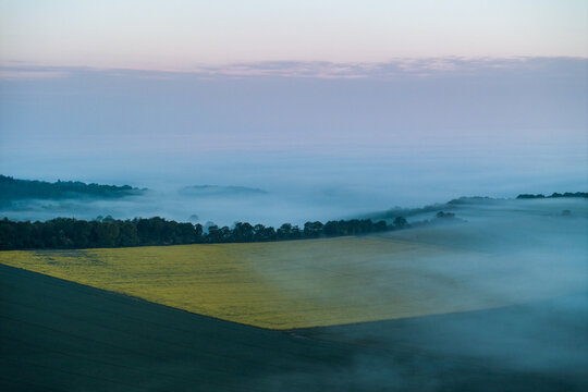 Aerial view of Nuffield countryside in the Chiltern Hills with yellow rapeseed fields and rolling hills partially covered by thick morning fog Nuffield, Oxfordshire, United Kingdom.