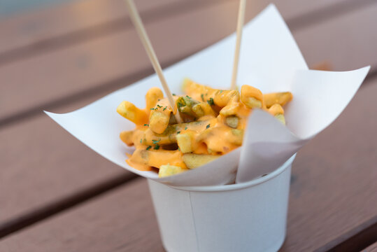 Crispy french fries topped with creamy cheese sauce served in paper cup with wooden sticks for tasty street food snack experience
