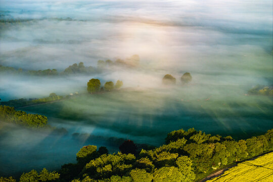 Aerial view of thick morning mist rolling over green fields and lush trees with soft sunlight filtering through the fog near Nuffield, Oxfordshire, United Kingdom.