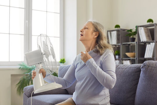 Senior elder woman sitting indoors, blowing fan feeling hot suffering from heat, uncomfortable, experiencing room summer discomfort, trying to cool down with fan, wishing for cooler temperature