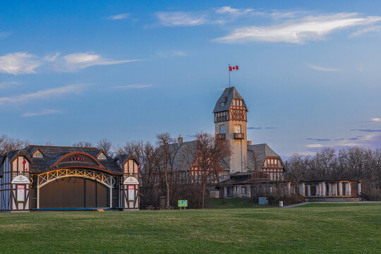 Historic Assiniboine Park Pavilion in Winnipeg during sunset with Canadian flag, theater stage and open green lawn under colorful evening sky.