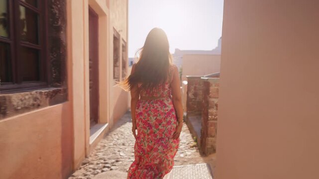 Young woman wearing colorful floral maxi dress runs through narrow cobblestone alley between traditional buildings during golden hour with sun flare and blue dome church visible. Santorini, Greece