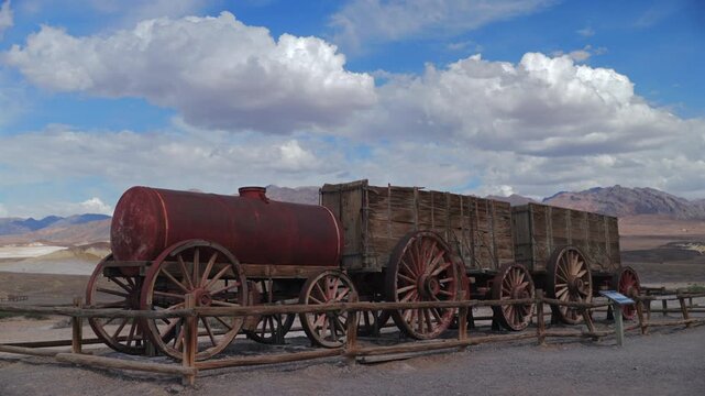 Vintage twenty mule team borax wagons with rustic red water tank and wooden ore carriers displayed behind wooden fence at Death Valley National Park under dramatic clouds.