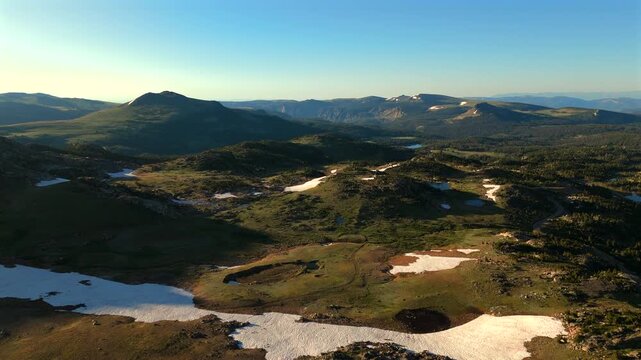 Stunning aerial footage of Beartooth Highway in Montana showcasing alpine meadows with melting snow patches and rolling mountain ranges under golden hour light. Scenic wilderness landscape.