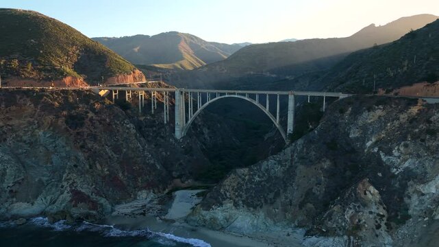 Aerial arch bridge at sunset glow, iconic concrete span framed by rugged canyon and pacific surf, warm backlight casting long shadows, cinematic composition for architecture, travel, coastal