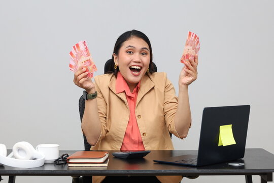 Shocked and amazed female employee holding a bundle of large amount of rupiah cash banknotes, working in the office at the desk with a laptop PC, business career achievement concept.