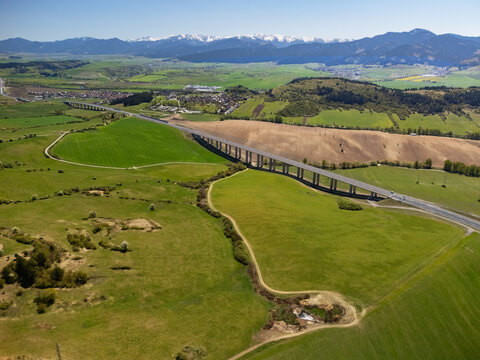 Aerial view of the D1 motorway viaduct crossing green fields and rolling hills with snow-capped mountains in the distance Likavka, Zilina Region, Slovakia.
